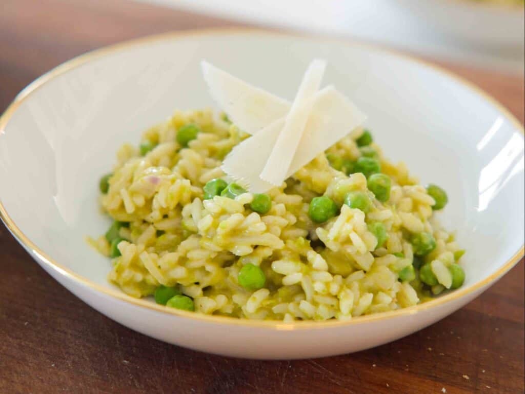 A bowl of creamy green pea risotto with green peas, topped with three thin shavings of Parmesan cheese, served in a white bowl with a gold rim on a wooden surface.